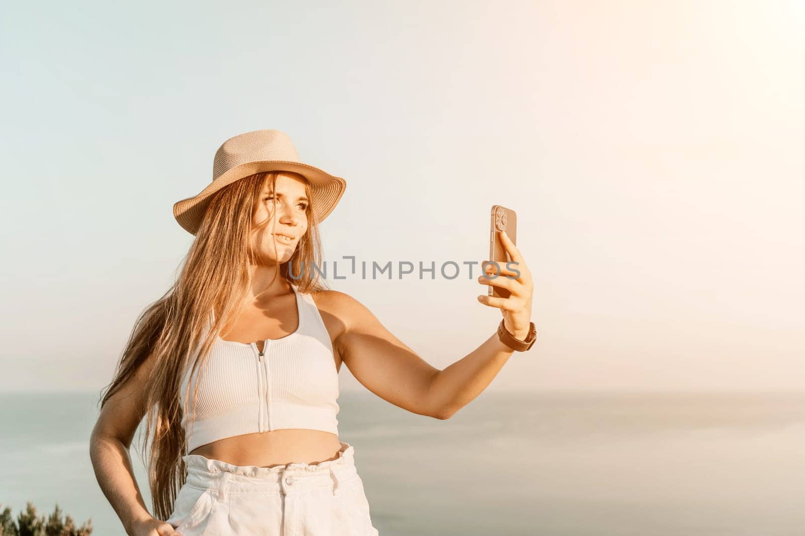 Selfie woman in a hat, white tank top, and shorts captures a selfie shot with her mobile phone against the backdrop of a serene beach and blue sea