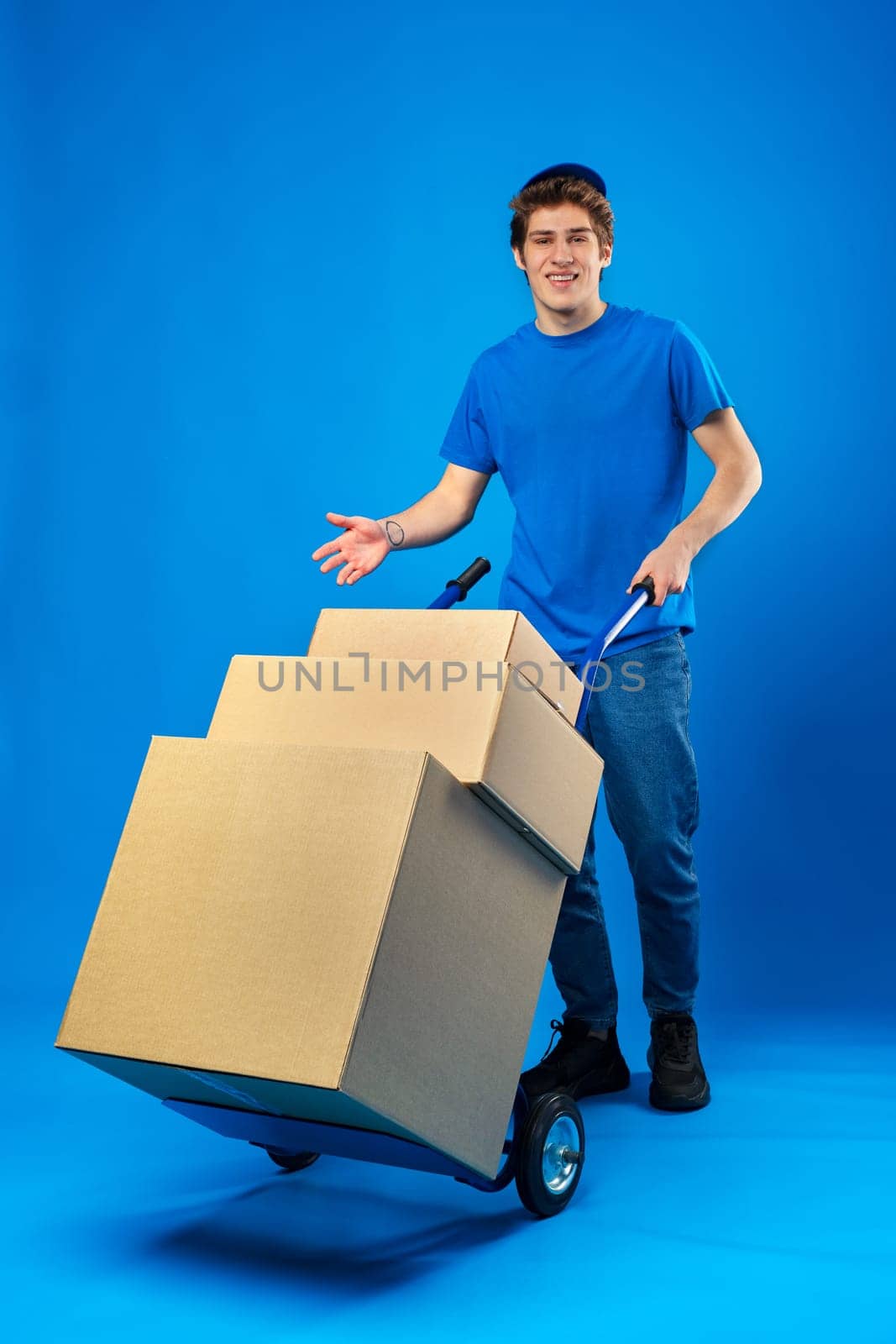 Young happy delivery man carrying boxes on truck against blue background in studio