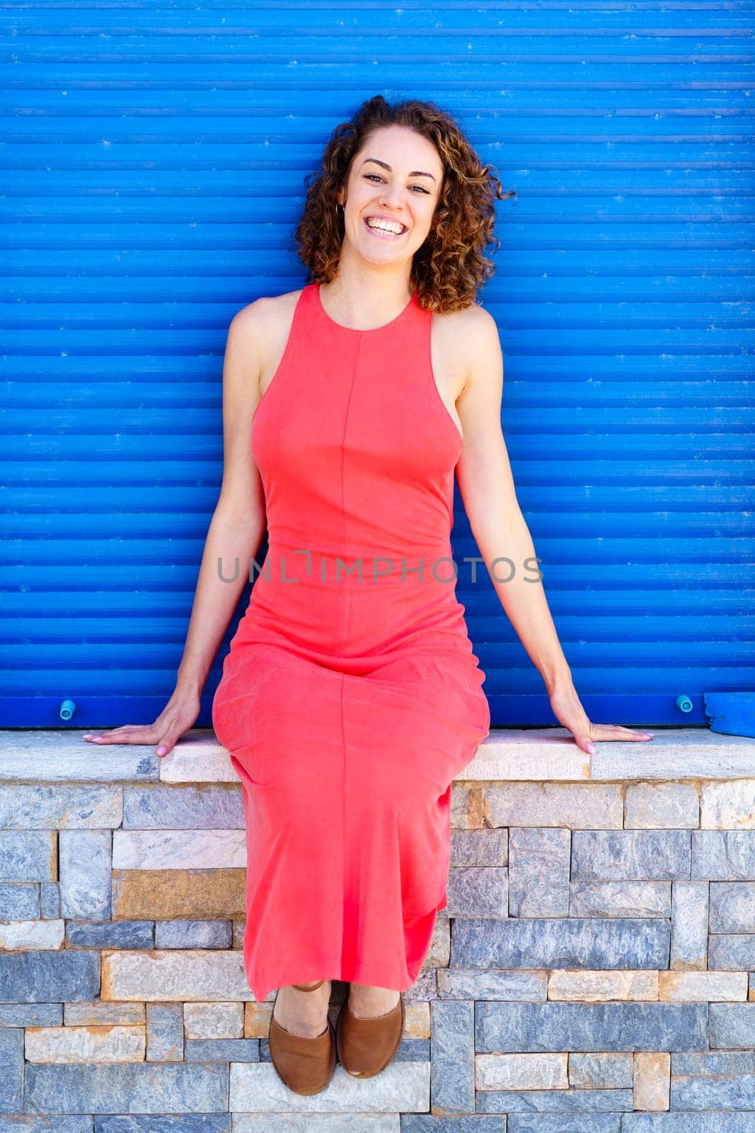 Cheerful young female in sleeveless red sundress and sandals looking at camera while relaxing on stone wall against blue shutters in daylight