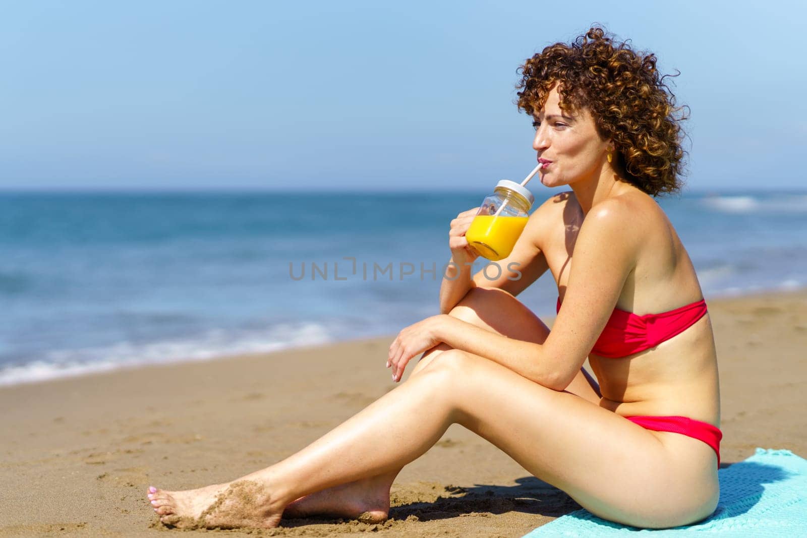 Side view of redhead woman in bikini sitting on beach of ocean in sunlight and drinking orange juice with straw from glass cup