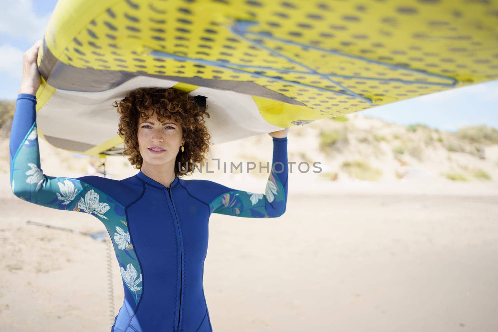 Positive curly haired female in trendy wetsuit looking at camera while lifting yellow SUP board, and standing on beach with sand dunes under blue sky on sunny summer day