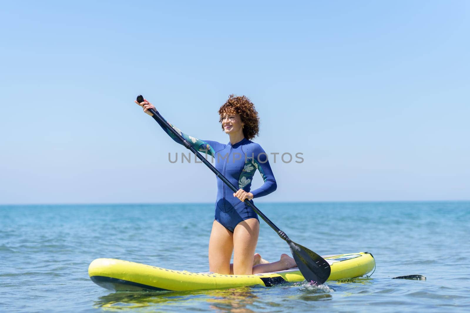 Full body view of young female with curly hair in wetsuit kneeling on paddleboard on blue sea while paddling and looking away