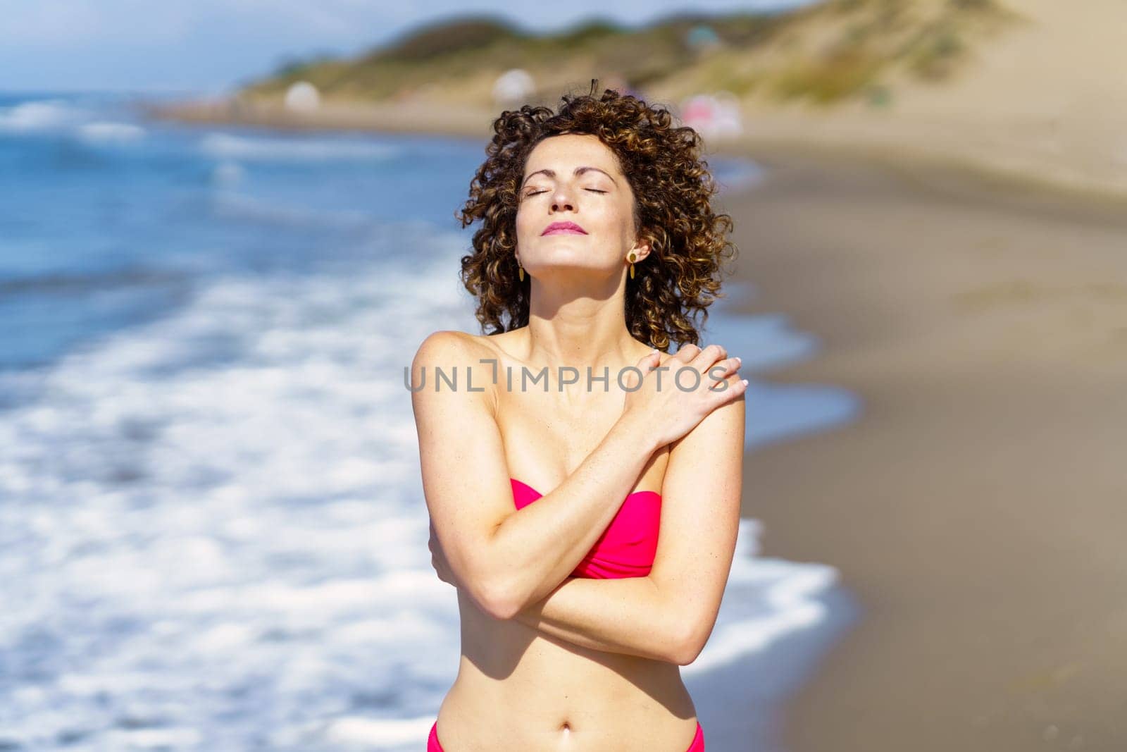 Young sensual female with curly ginger hair with hands crossed and touching bare shoulder, while facing sun with eyes closed in daylight and standing in blurred sea beach