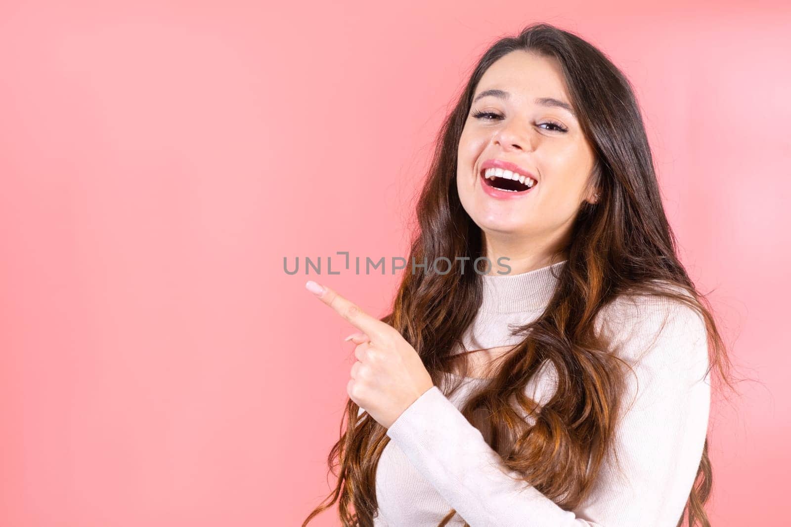 Long-haired woman with shocked and amused expression points finger to new visitor in pink studio. Lady smiles covering mouth at pink studio wall