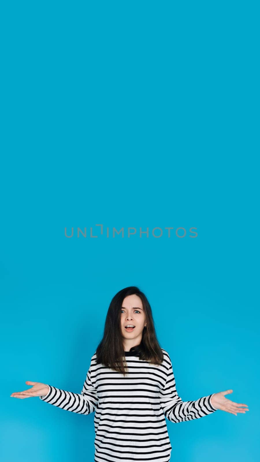 Captivating Portrait of Astonished Woman in Striped Pullover - Surprised and Amazed Female Model with Arms Extended - Isolated on Blue Background - Perfect for Expressing Shock, Surprise, and Emotion.