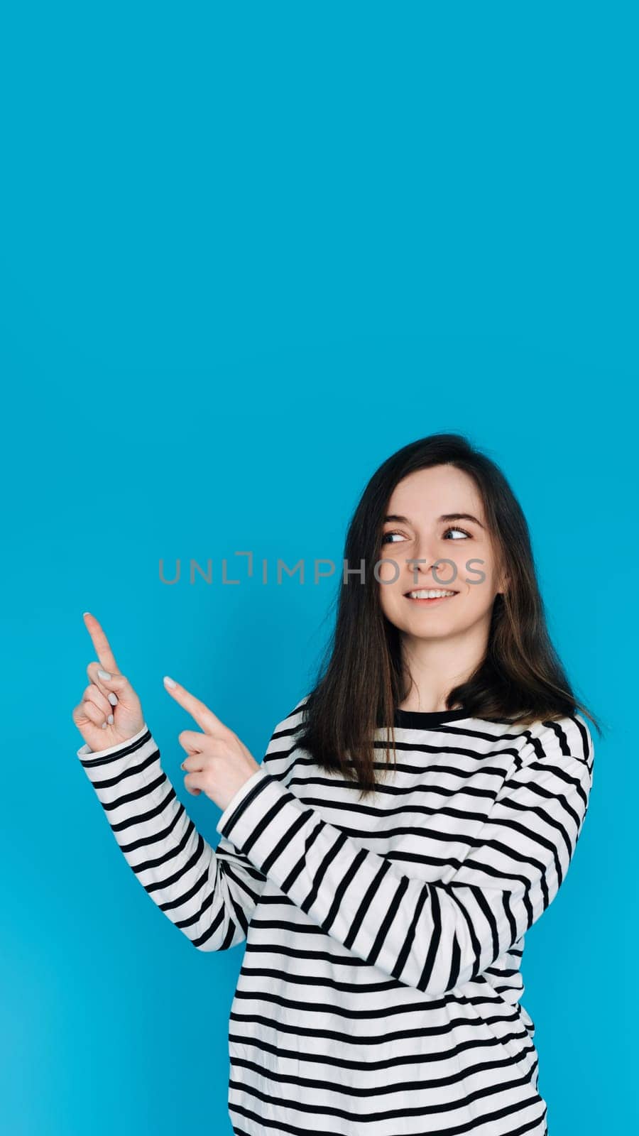 Adorable Girl in Striped Pullover Pointing and Gazing at Empty Space - Charming Young Female Model with Expressive Gestures - Isolated on Blue Background - Perfect for Conceptual, Advertising by ViShark