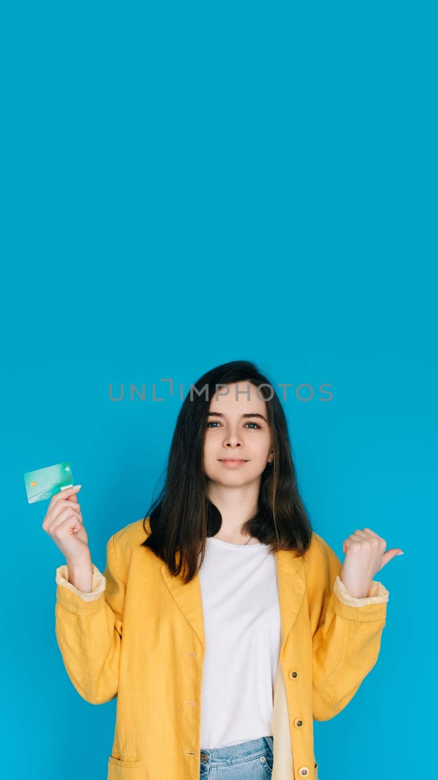 Happy Woman Pointing Thumb at Empty Space with Credit Card - Positive and Attractive Female Expressing Satisfaction and Financial Confidence, Isolated on Blue Background - Ideal for Advertising