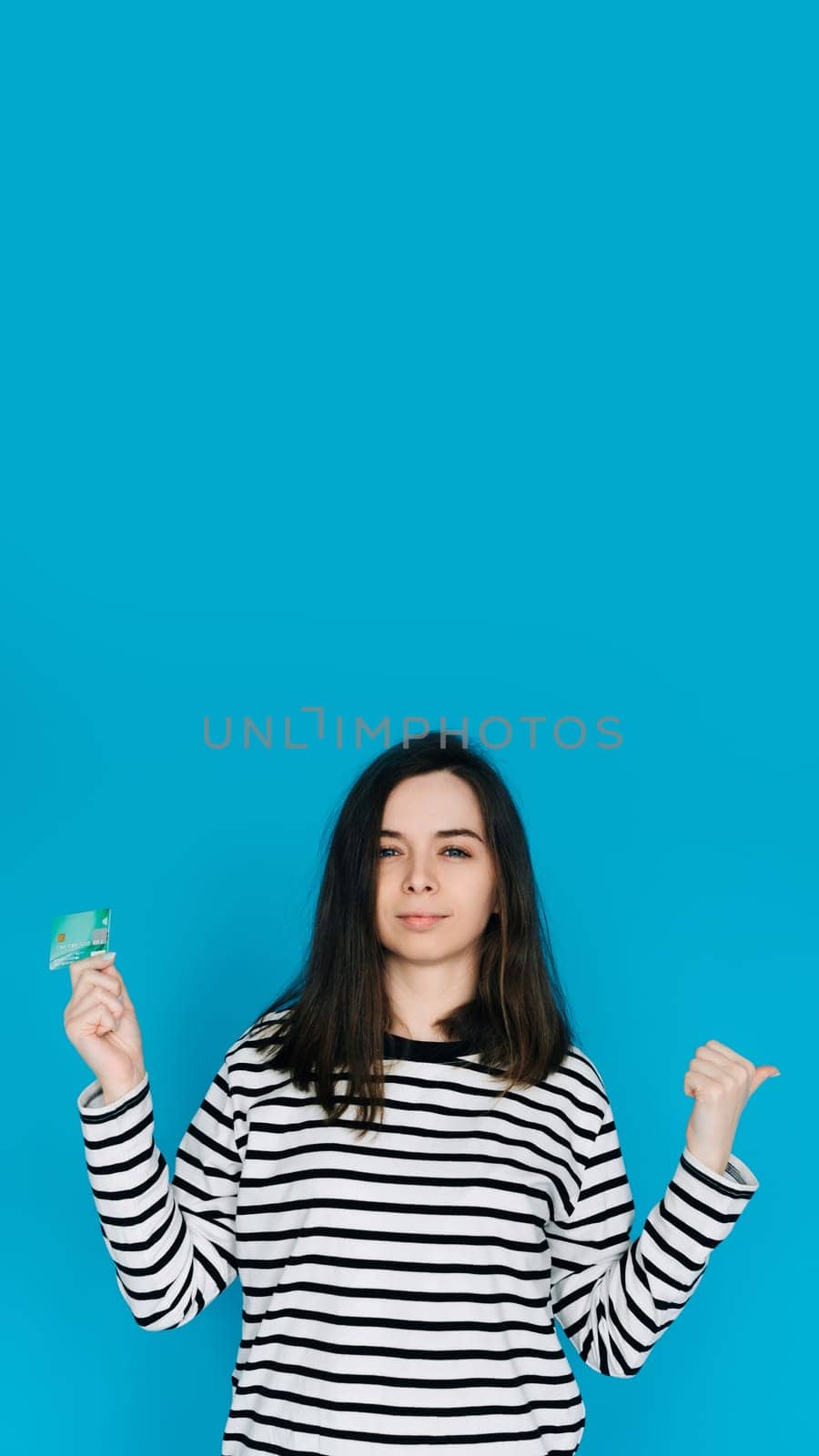 Woman Holding Credit Card, Pointing with Thumb in Empty Space - Female Expressing Confidence and Financial Freedom - Blue Background - Perfect for Banking, E-commerce, and Financial Services Concepts.