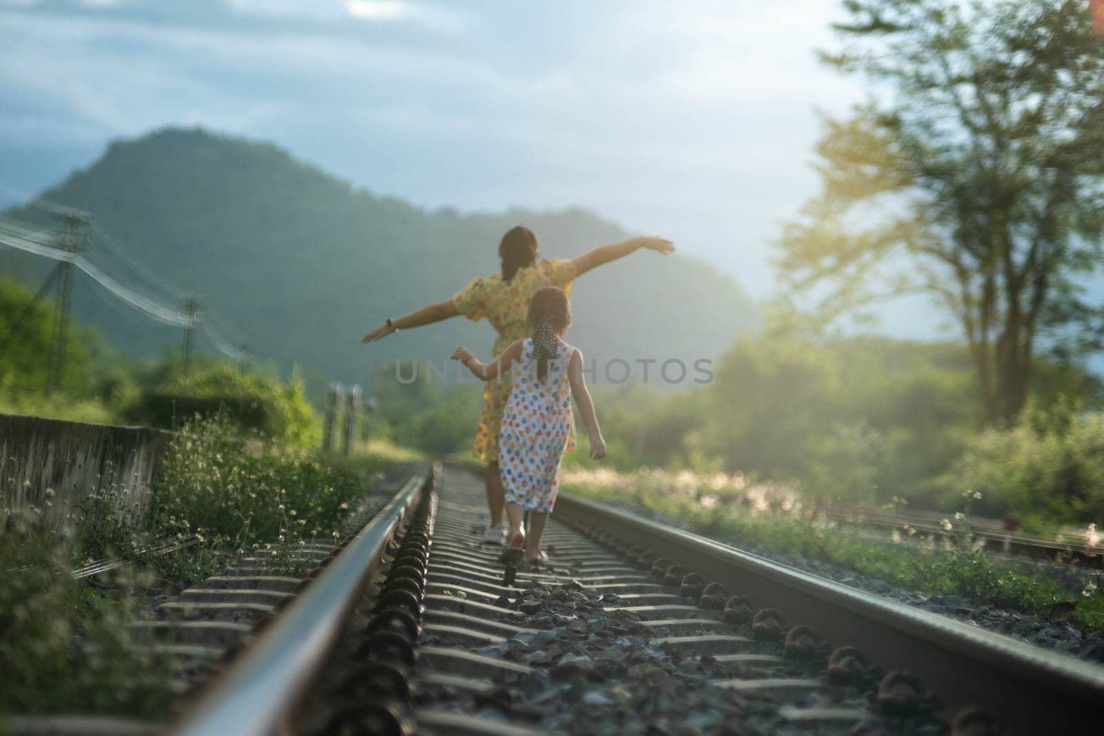 Mother and cute daughter walking on the railroad in the daytime. Happy family walking on the railway against the background of mountains and greenery. by TEERASAK