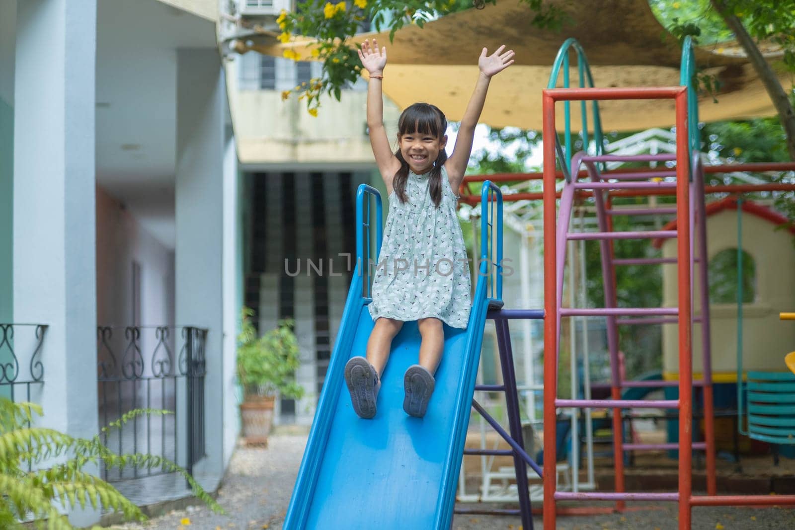 Happy girl playing on the slide. Happy little asian girl sliding and playing at outdoor playground in park on summer vacation. Healthy activity. by TEERASAK