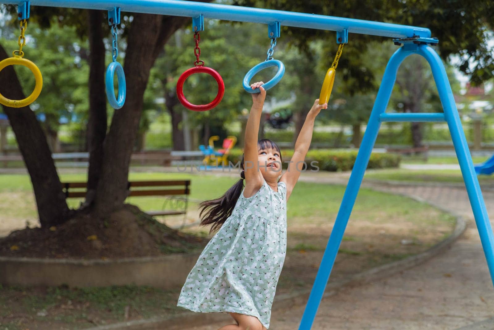Happy girl hanging on monkey bar by hand doing exercise. Little Asian girl playing at outdoor playground in the park on summer vacation. Healthy activity. by TEERASAK