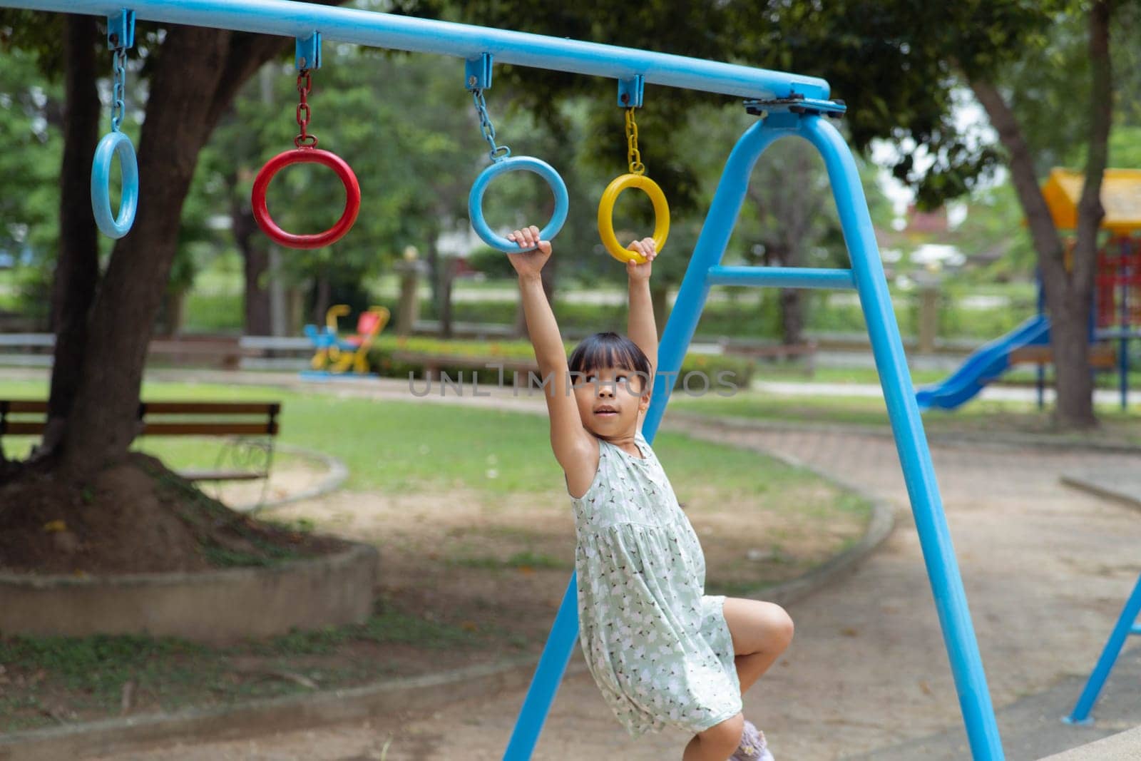 Happy girl hanging on monkey bar by hand doing exercise. Little Asian girl playing at outdoor playground in the park on summer vacation. Healthy activity. by TEERASAK