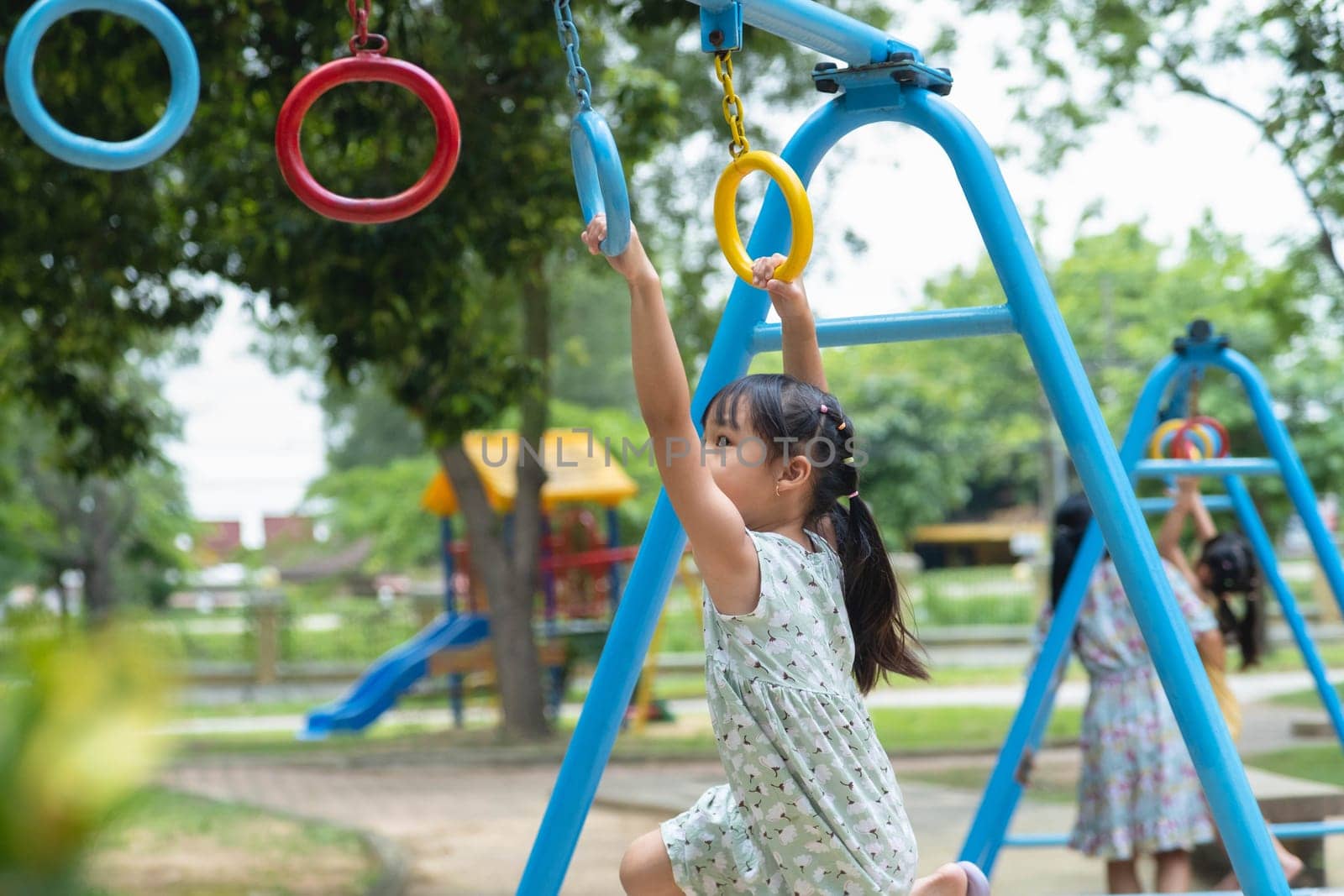 Happy girl hanging on monkey bar by hand doing exercise. Little Asian girl playing at outdoor playground in the park on summer vacation. Healthy activity. by TEERASAK