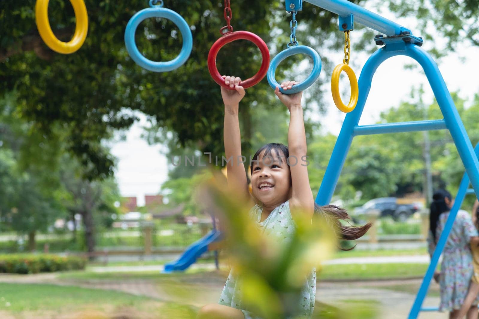 Happy girl hanging on monkey bar by hand doing exercise. Little Asian girl playing at outdoor playground in the park on summer vacation. Healthy activity.