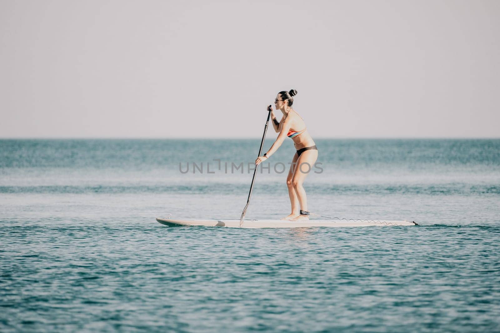 Sea woman sup. Silhouette of happy middle aged tanned woman in rainbow bikini, surfing on SUP board, confident paddling through water surface. Idyllic sunset. Active lifestyle at sea or river. by panophotograph