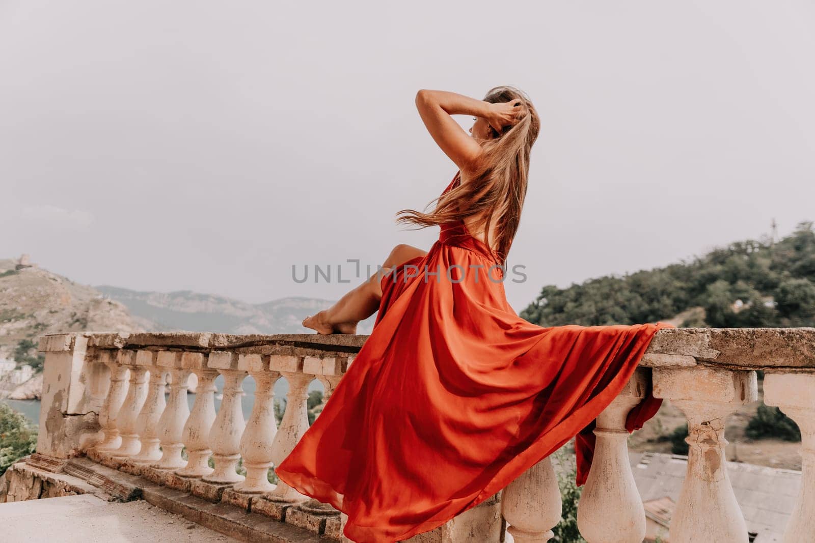 Side view a Young beautiful sensual woman in a red long dress posing on a volcanic rock high above the sea during sunset. Girl on the nature on overcast sky background. Fashion photo