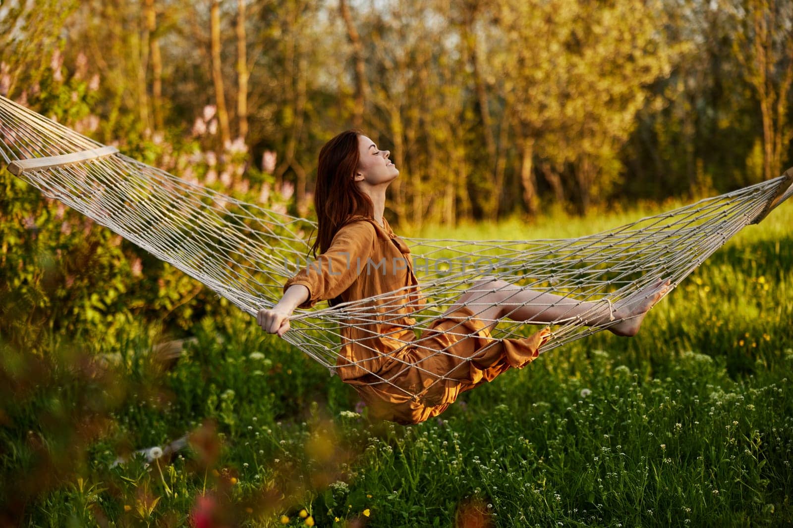 a happy woman in a long orange dress is resting sitting in a hammock at the dacha, smiling pleasantly looking away, illuminated by the summer sun during sunset. Horizontal photo. High quality photo