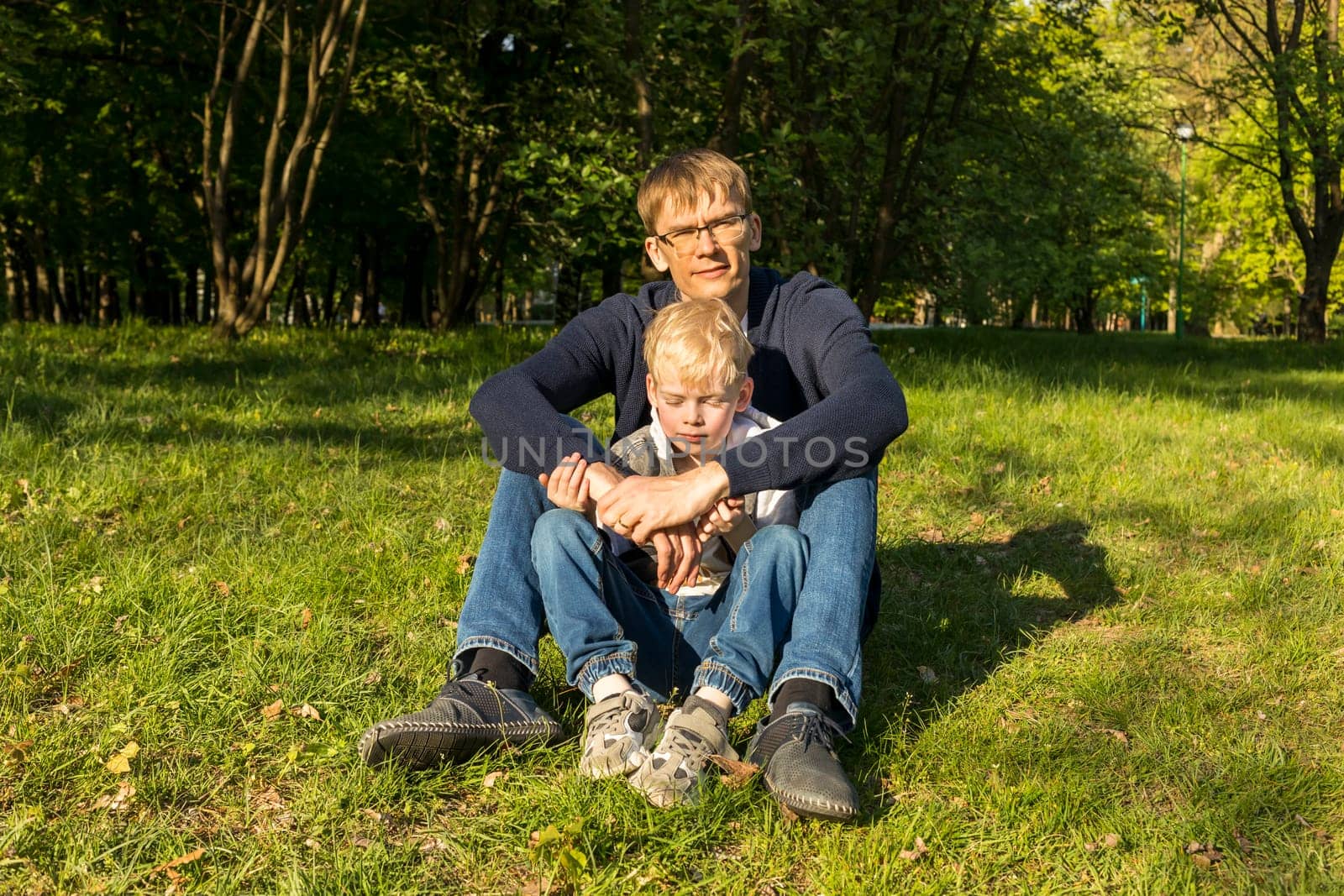 Father's Day. Father And Son Sit On Grass In Park, Enjoying Time Together. Summer Time. Happy Parenthood, Family Leisure Time, Emotional Connection, Love And Care. Horizontal Plane.