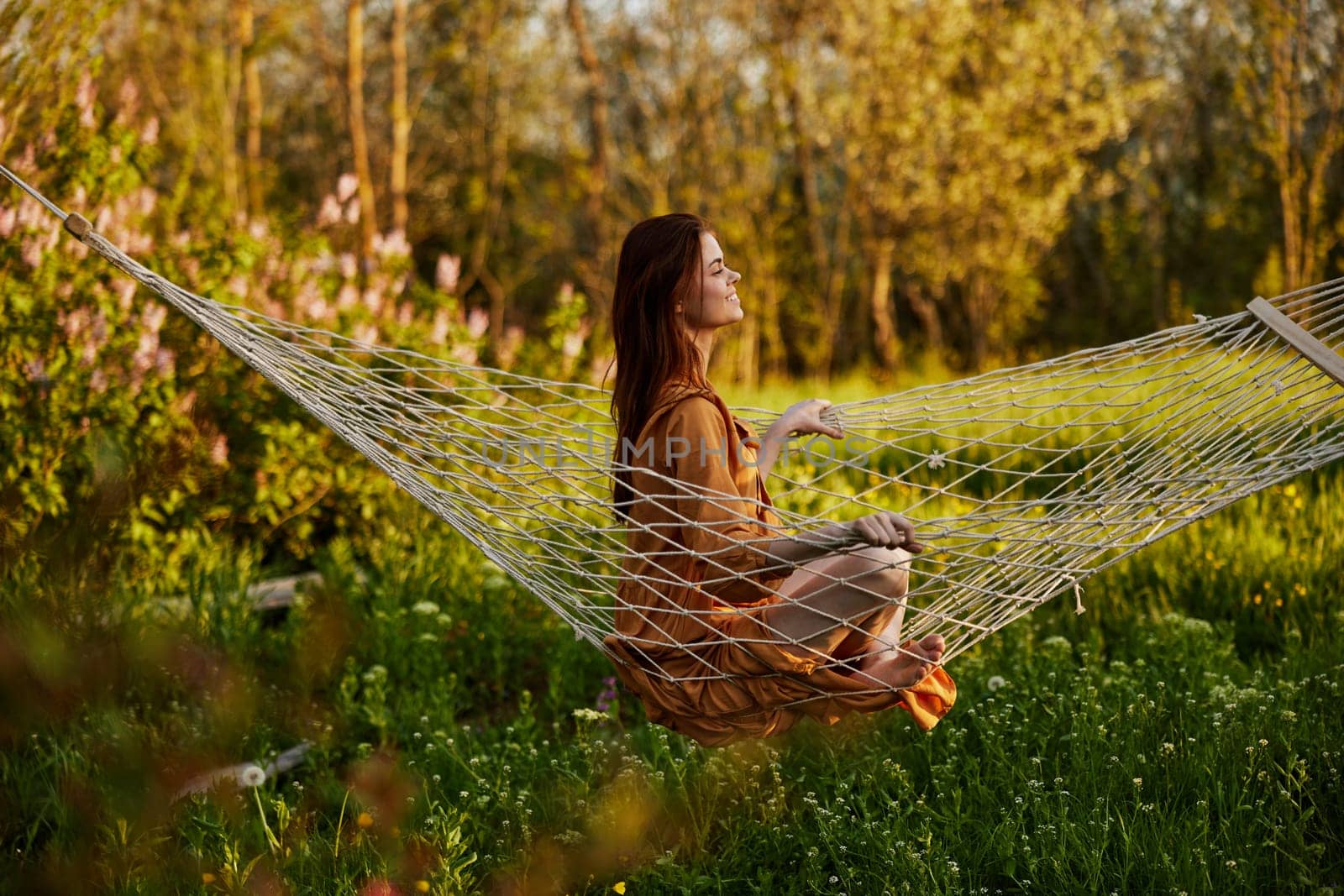 a happy woman in a long orange dress is resting sitting in a hammock at the dacha, smiling pleasantly looking away, illuminated by the summer sun during sunset. Horizontal photo by Vichizh