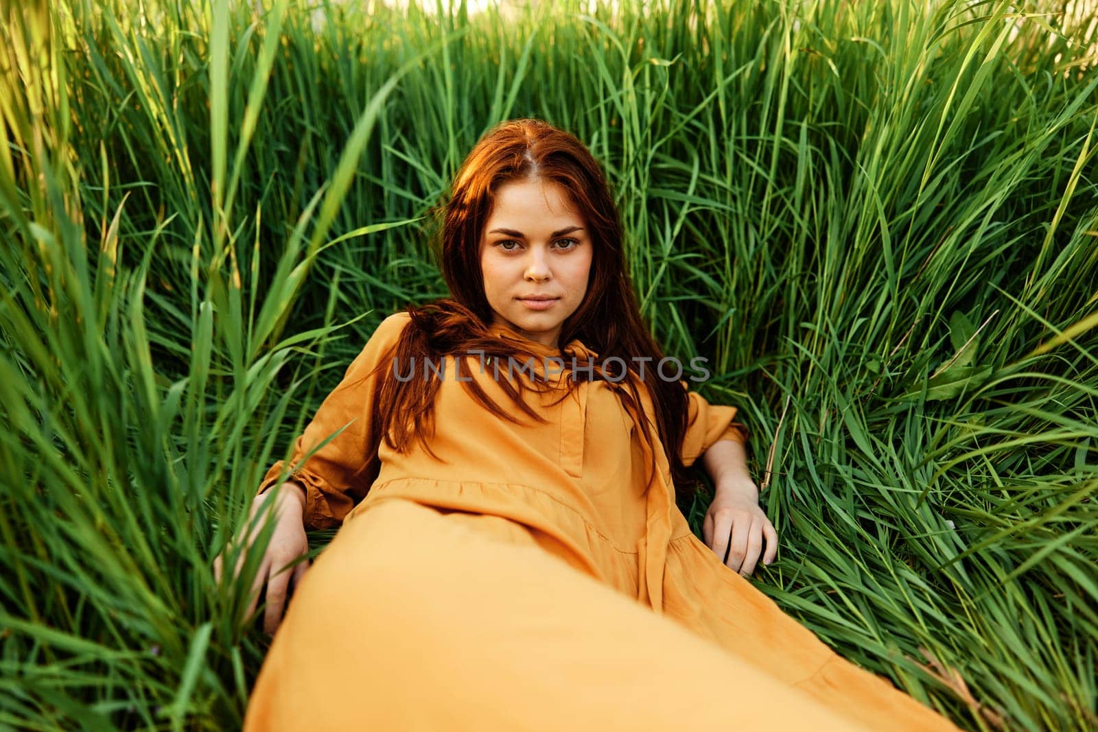 a happy woman lies in the tall green grass in a long orange dress and smiling pleasantly looks into the camera enjoying nature. Close horizontal photo. High quality photo