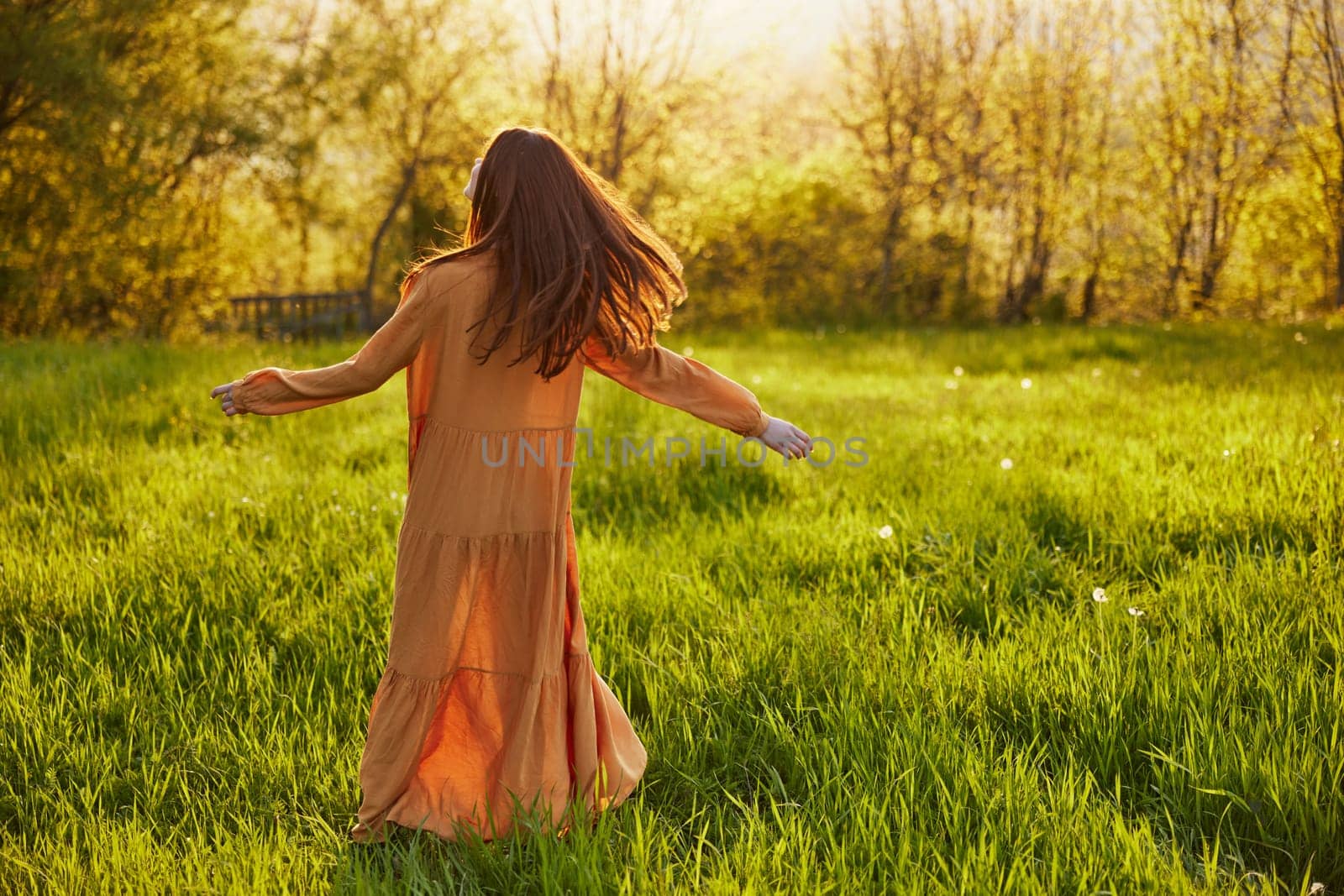 a relaxed slender woman enjoys the sunset standing in a green field with tall grass in an orange dress with her back to the camera, in warm summer weather. Horizontal photo. High quality photo