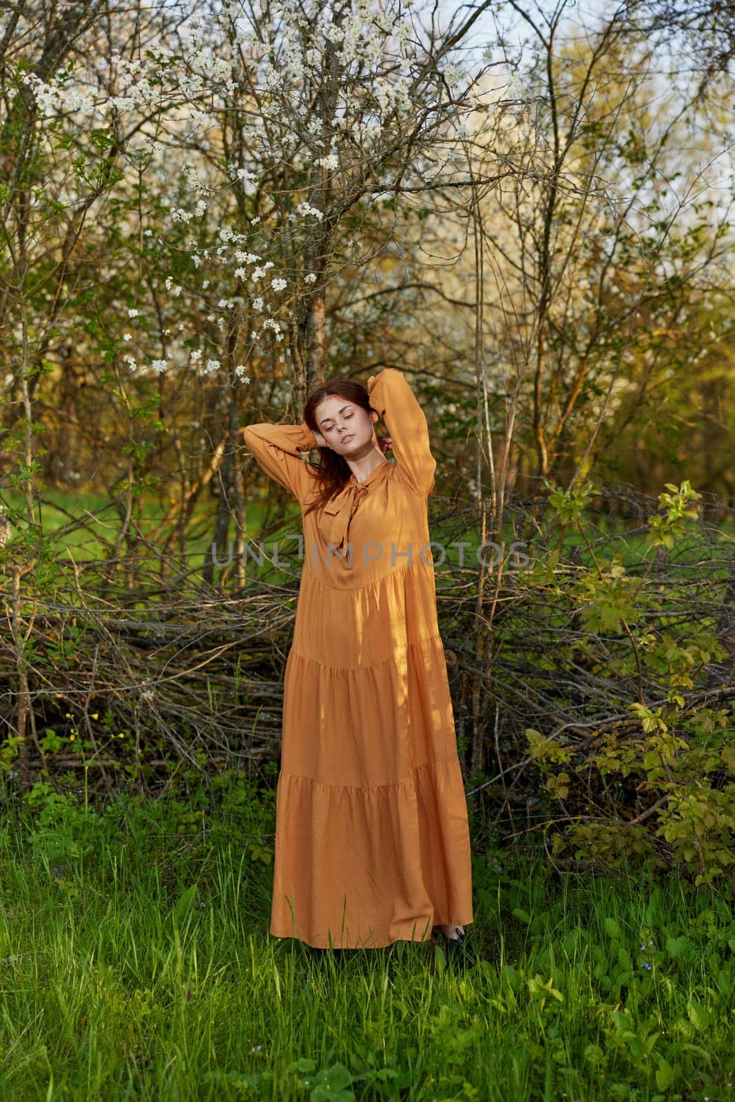an elegant, sophisticated woman poses standing near a wicker fence in a dacha in a long orange dress, straightening her hair with her hands by Vichizh