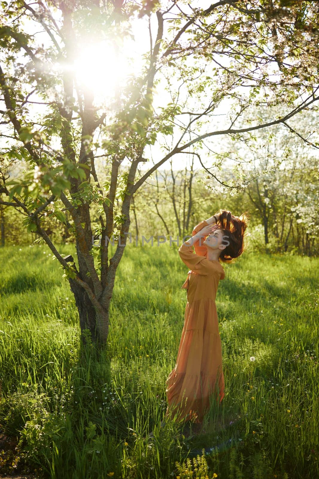 a beautiful, joyful woman stands in a long orange dress, in the countryside, near a tree blooming with white flowers, during sunset, illuminated from behind and touches her long hair by Vichizh