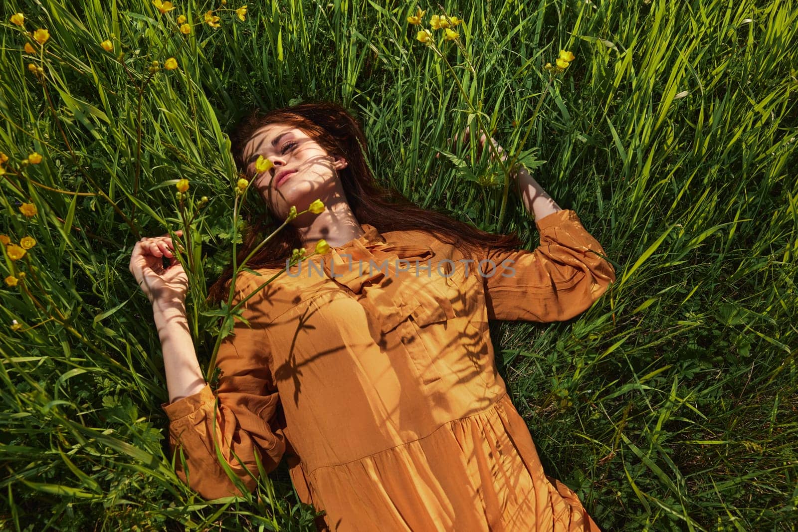 a calm woman with long red hair lies in a green field with yellow flowers, in an orange dress with her eyes closed, spreading her arms to the sides, enjoying peace and recuperating by Vichizh