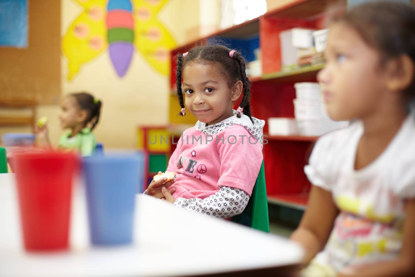 Child, classroom and lunch in portrait with smile for education at a table with children. Break, kids and learning at school with apple in kindergarten with chair or desk with food with friends. by YuriArcurs
