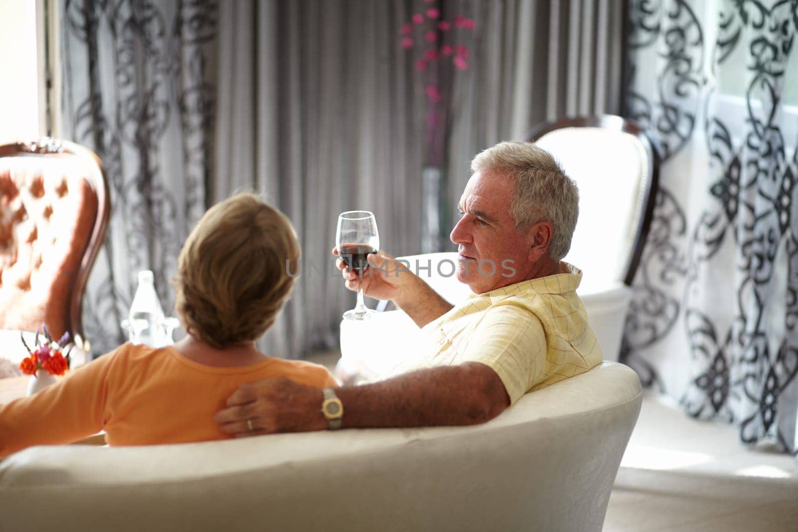 Retirement, sofa and an old couple drinking wine in their hotel room while on holiday or vacation together. Toast, love or relax with a senior man and woman bonding at a luxury resort for romance by YuriArcurs