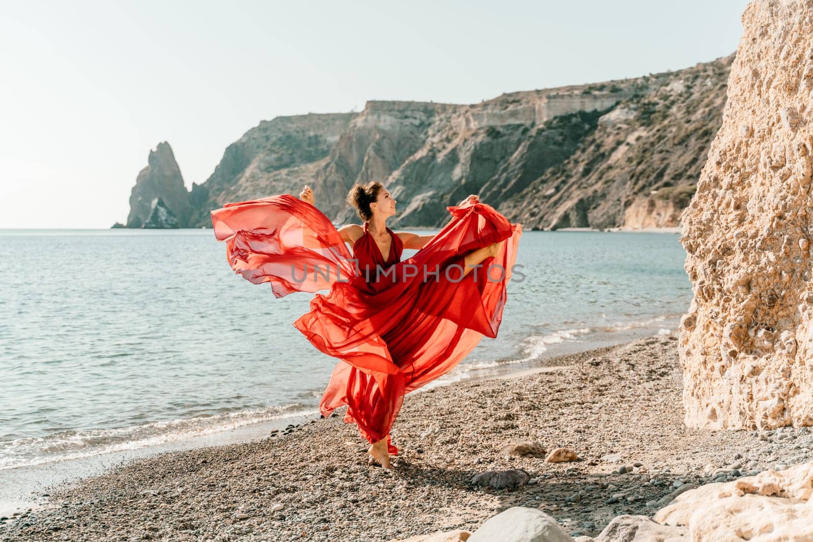 Woman red dress sea. Female dancer in a long red dress posing on a beach with rocks on sunny day. Girl on the nature on blue sky background. by Matiunina