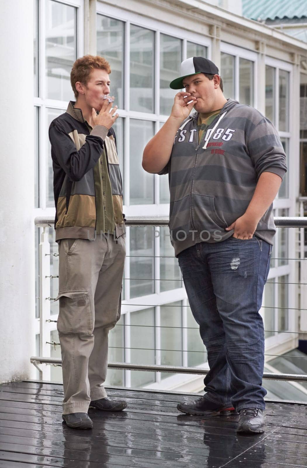 Teen men, college friends and smoking outside education and school building outdoor. Rebel, guys and teenager students with tobacco and cigarette smoke in rain with a male young friend of university.