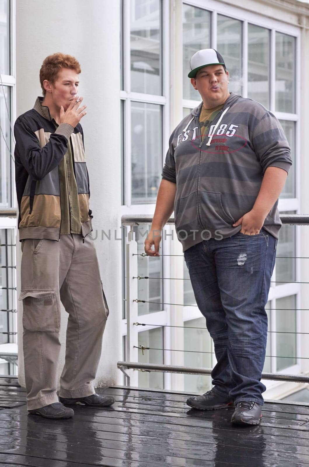 Teen men, friends and smoking outside education and school building outdoor. Rebel, guys and teenager students with tobacco and cigarette smoke in the rain with a male young friend of university.