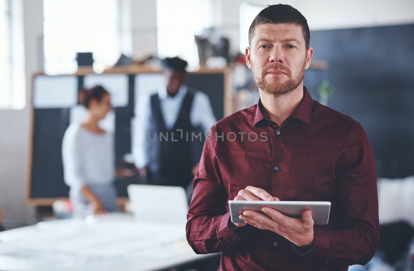 Its the smart way to do business. Cropped portrait of a handsome young businessman using a tablet in the office with his colleagues in the background. by YuriArcurs