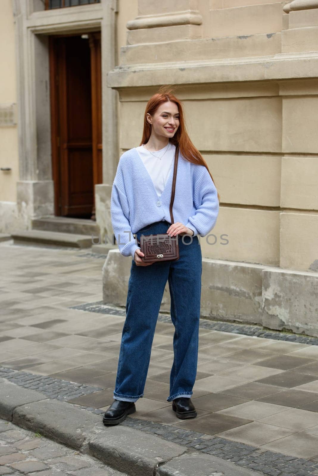 a red-haired girl in a blue jeans and a sweater poses outside with a small leather handbag.