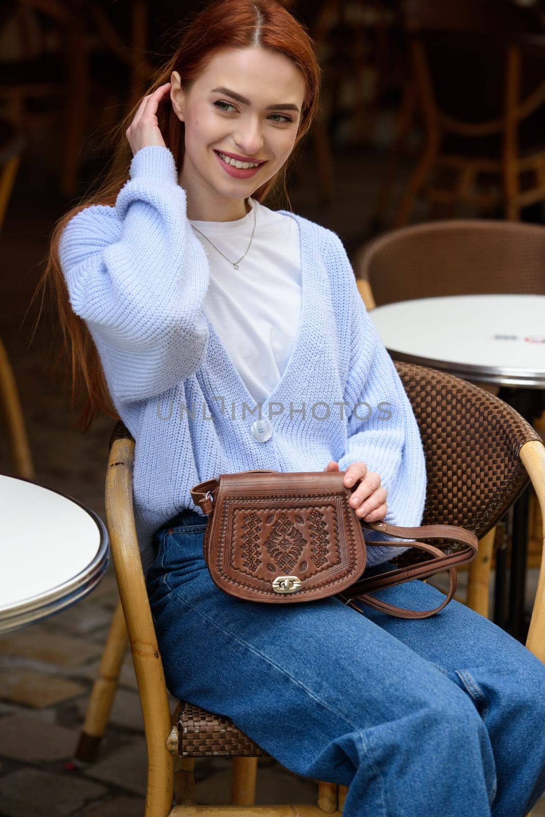 a red-haired girl in a blue jeans and a sweater poses outside with a small leather handbag.