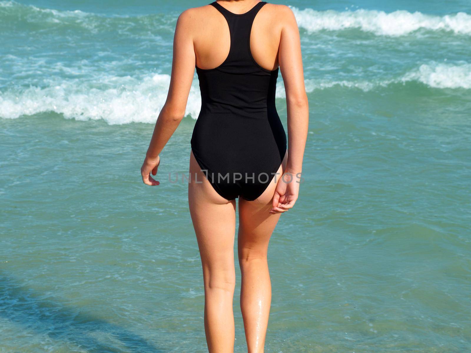 girl walks into the sea in a black swimsuit back view by Annado