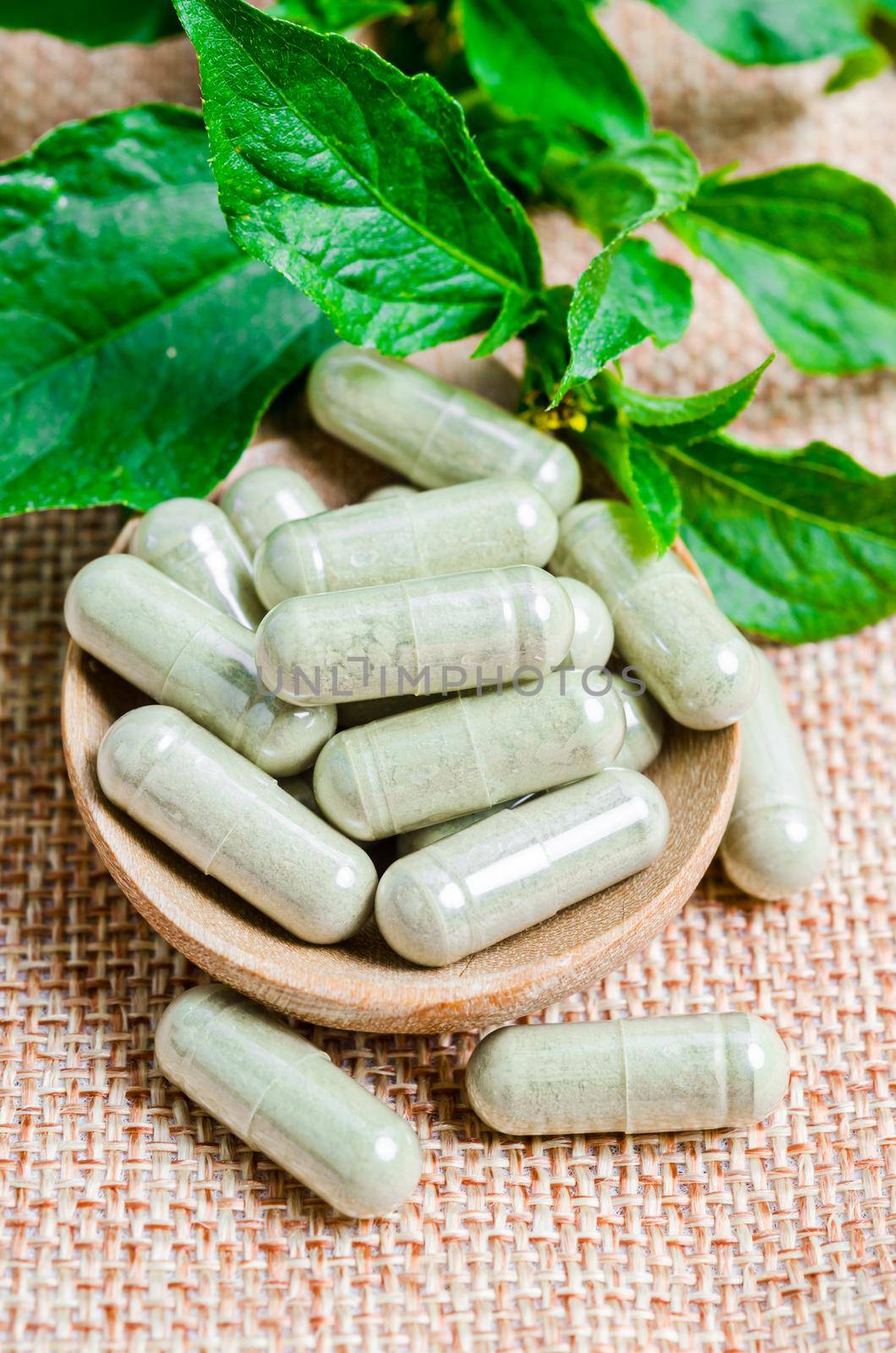 Pile of herbal medicine capsules in wooden spoon with green leaf on sack background.