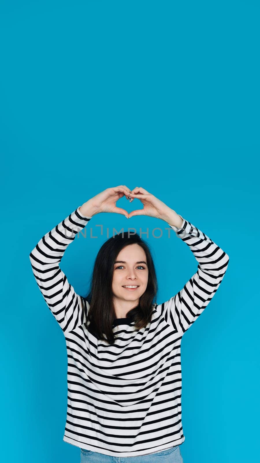 Happy Woman in Striped Sweater Spreading Love and Positivity - Smiling Girl with Raised Arms Forming Heart Shape, Expressing Joy and Affection - Isolated Blue Background - Ideal for Happiness, Love,