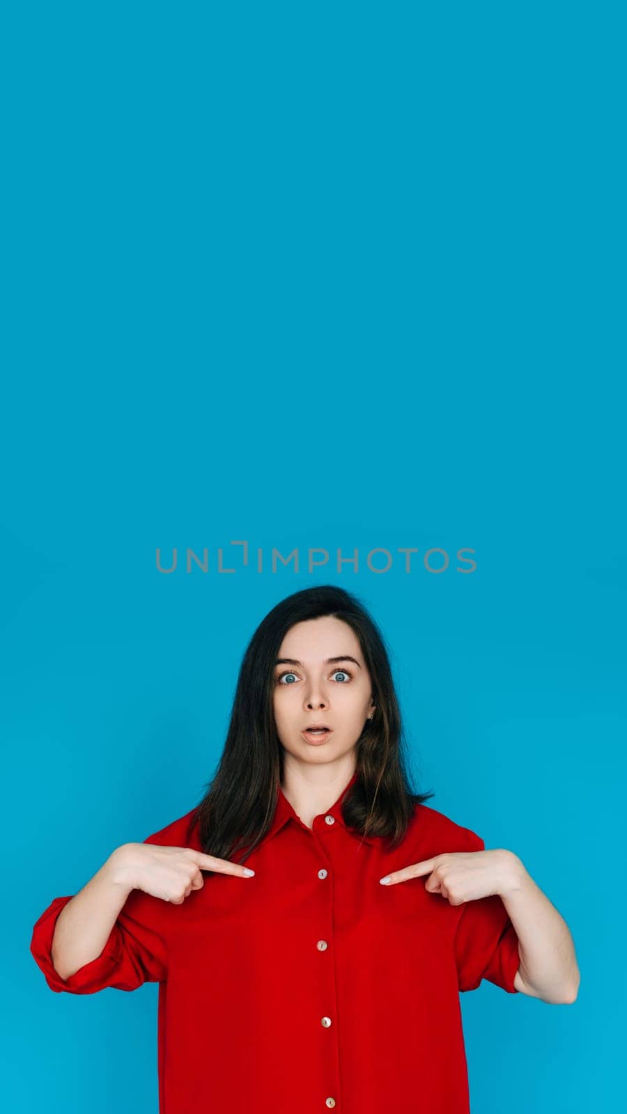 A photo of a lovely impressed young woman in a red shirt, surprised face, pointing her finger, isolated on a blue background - Perfect for Emphasizing Surprise and Amazement.