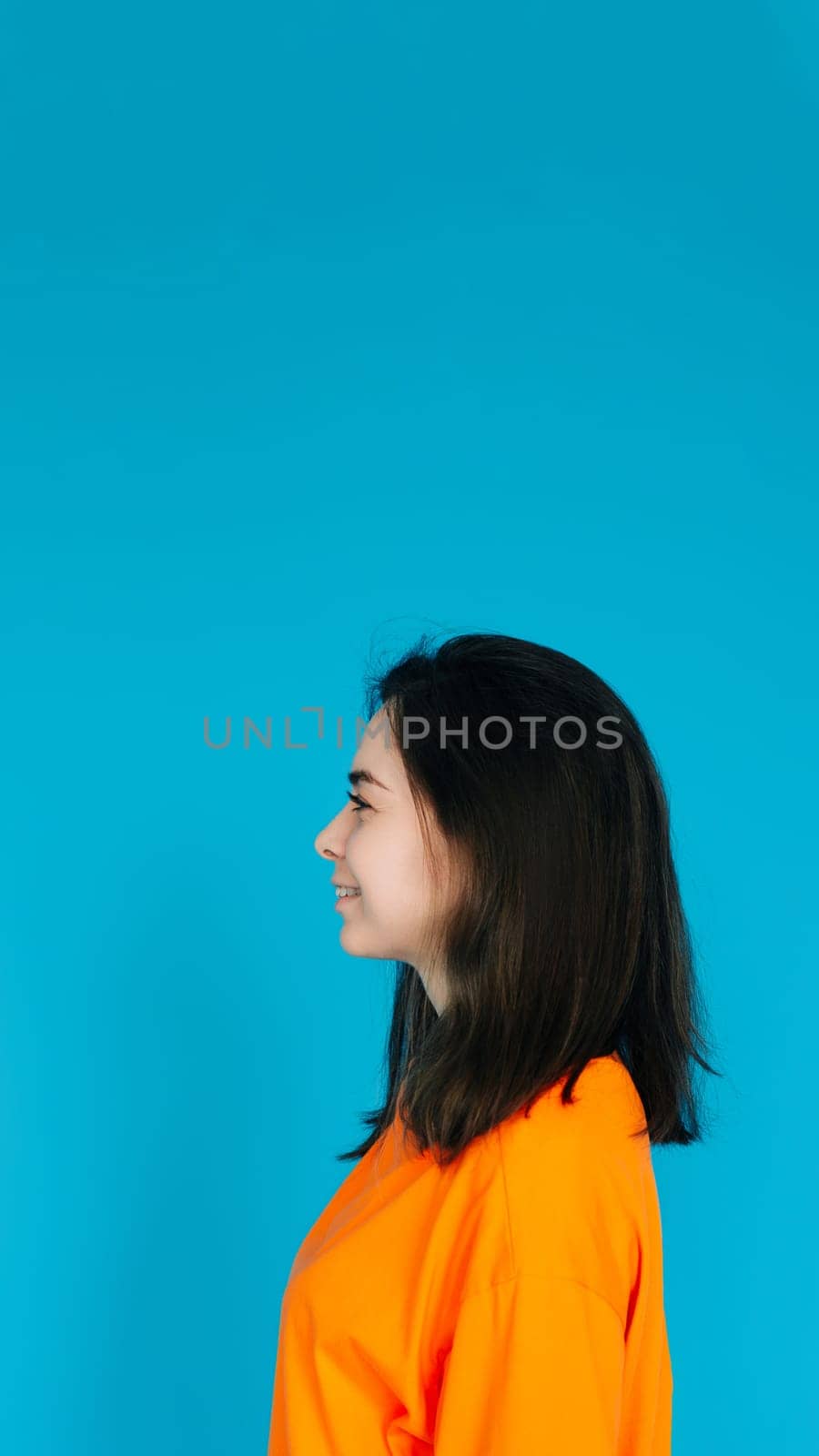 Radiant Young Woman in Stylish Orange Attire Smiling with a Beaming Toothed Smile, Profile Portrait, Isolated on Blue Background by ViShark