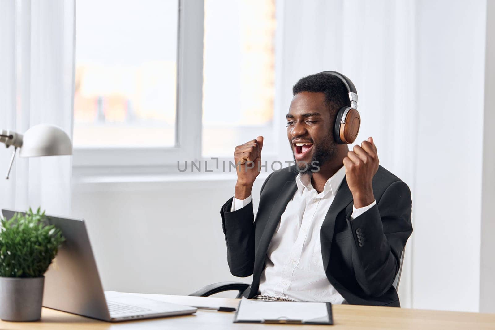 An African-American man sits at his desk in front of his laptop, wearing headphones and chatting on a video call, listening to music. The concept of student business training and online work. by SHOTPRIME