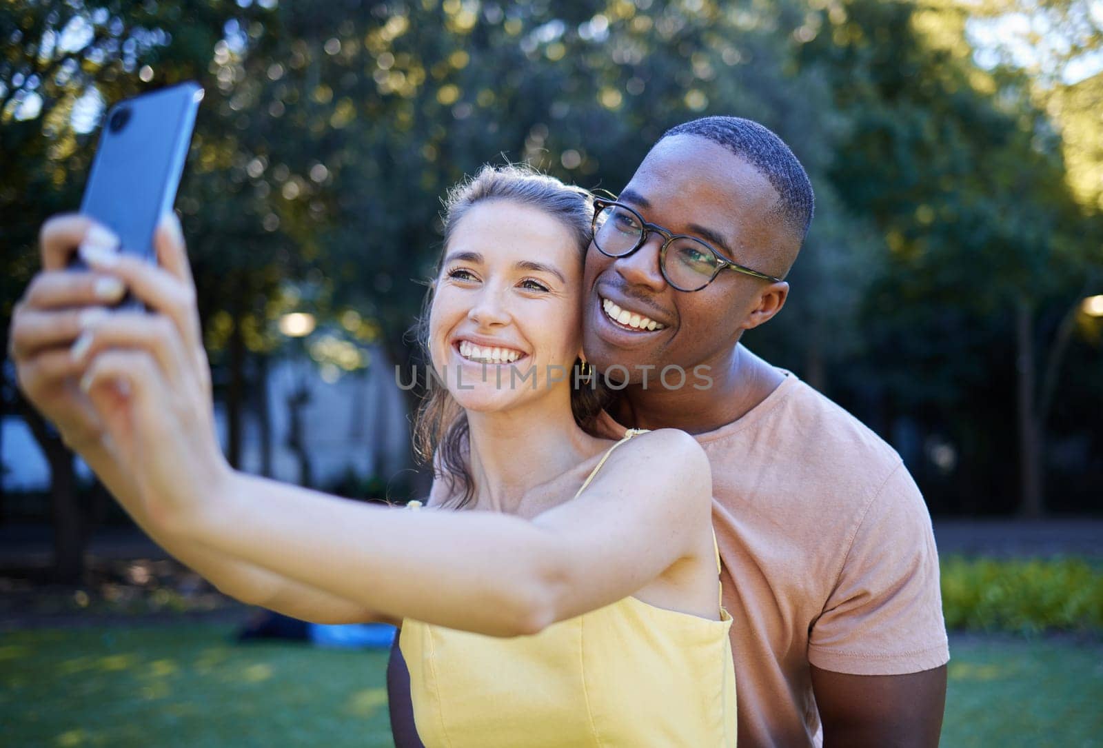 Selfie, phone and interracial couple smile in park enjoying weekend, summer holiday and quality time together. Nature, dating and happy man and woman relax, calm and hug to take picture on smartphone by YuriArcurs