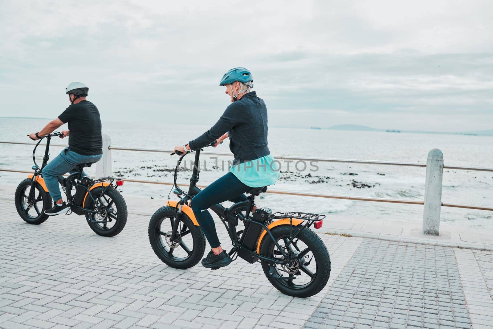 Couple, helmet or electrical bike by sea or ocean beach in bonding transport, clean energy or sustainability travel. Technology, electric or eco friendly bicycle for happy woman or mature cycling man by YuriArcurs