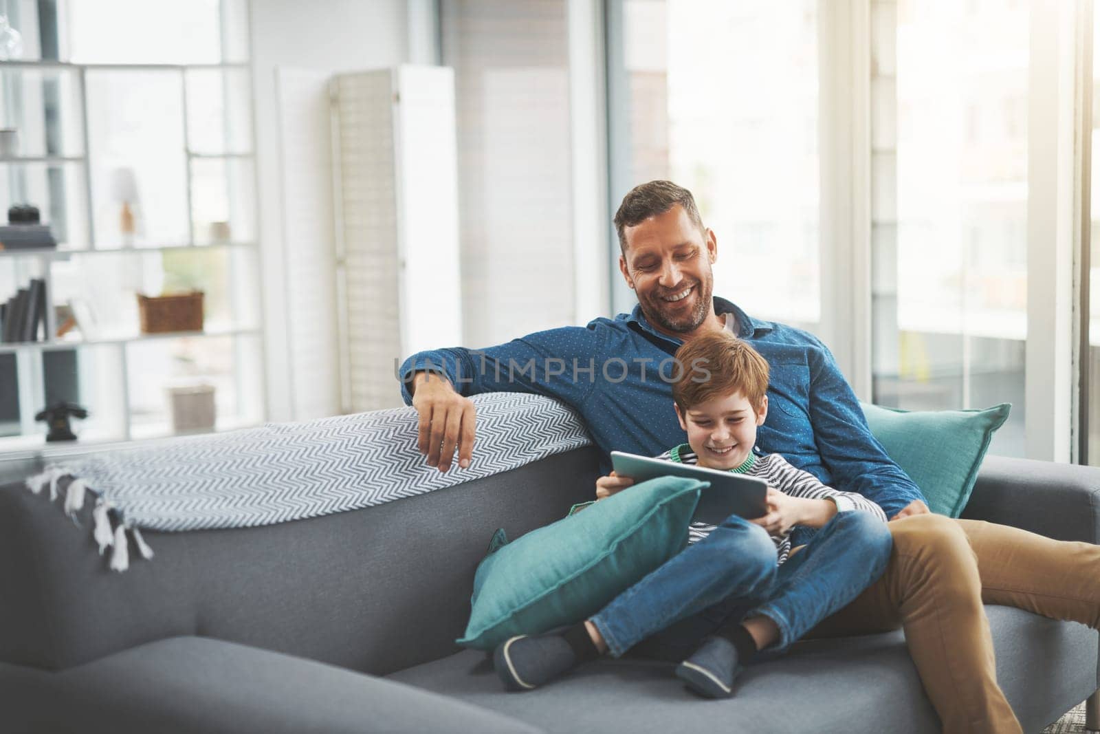 Learning in a fun way. a cheerful little boy browsing on a digital tablet while lying on his fathers lap at home during the day. by YuriArcurs