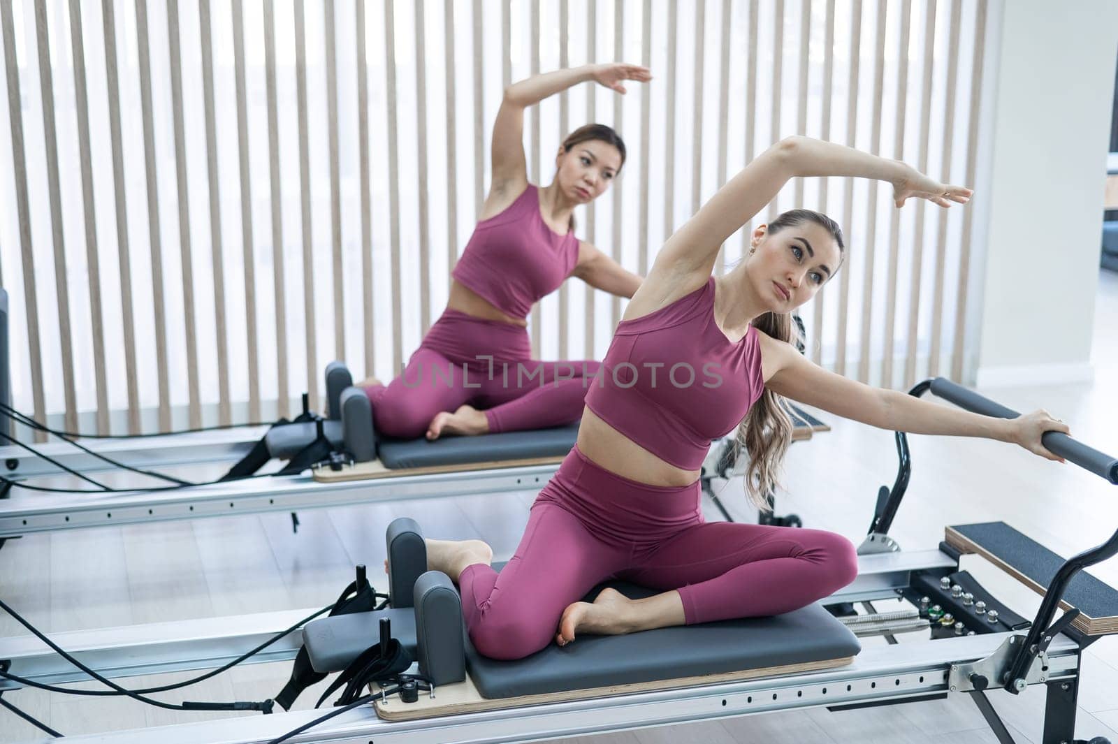 Two Asian women in pink sportswear doing side bends on a reformer machine. Pilates classes