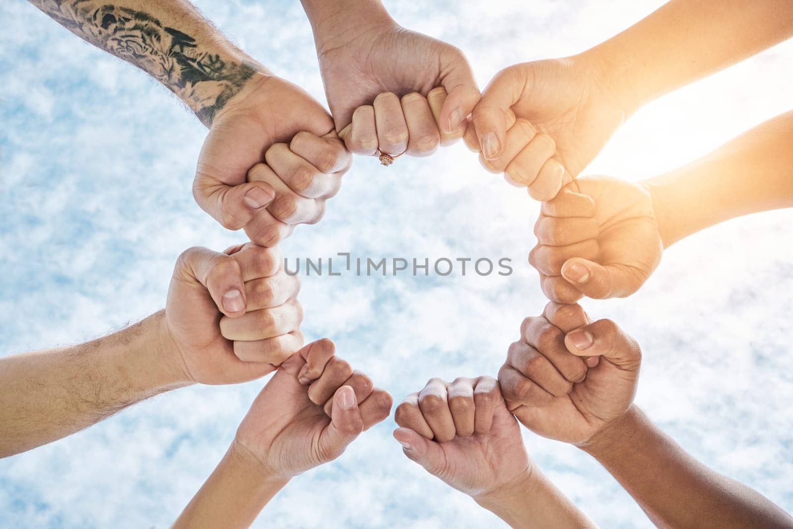 Hands in circle, fist and blue sky, community in collaboration for support and diversity together from below. Teamwork, power hand sign and sunshine, positive group of people in solidarity and huddle.