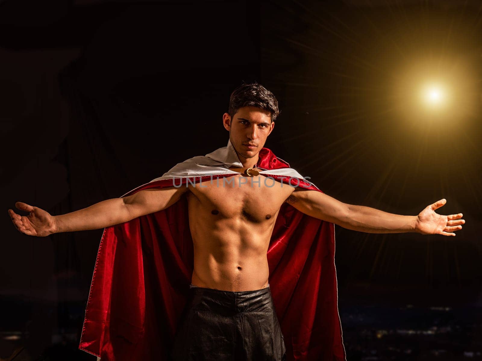 Handsome muscular shirtless young man standing confident, in cape against black background