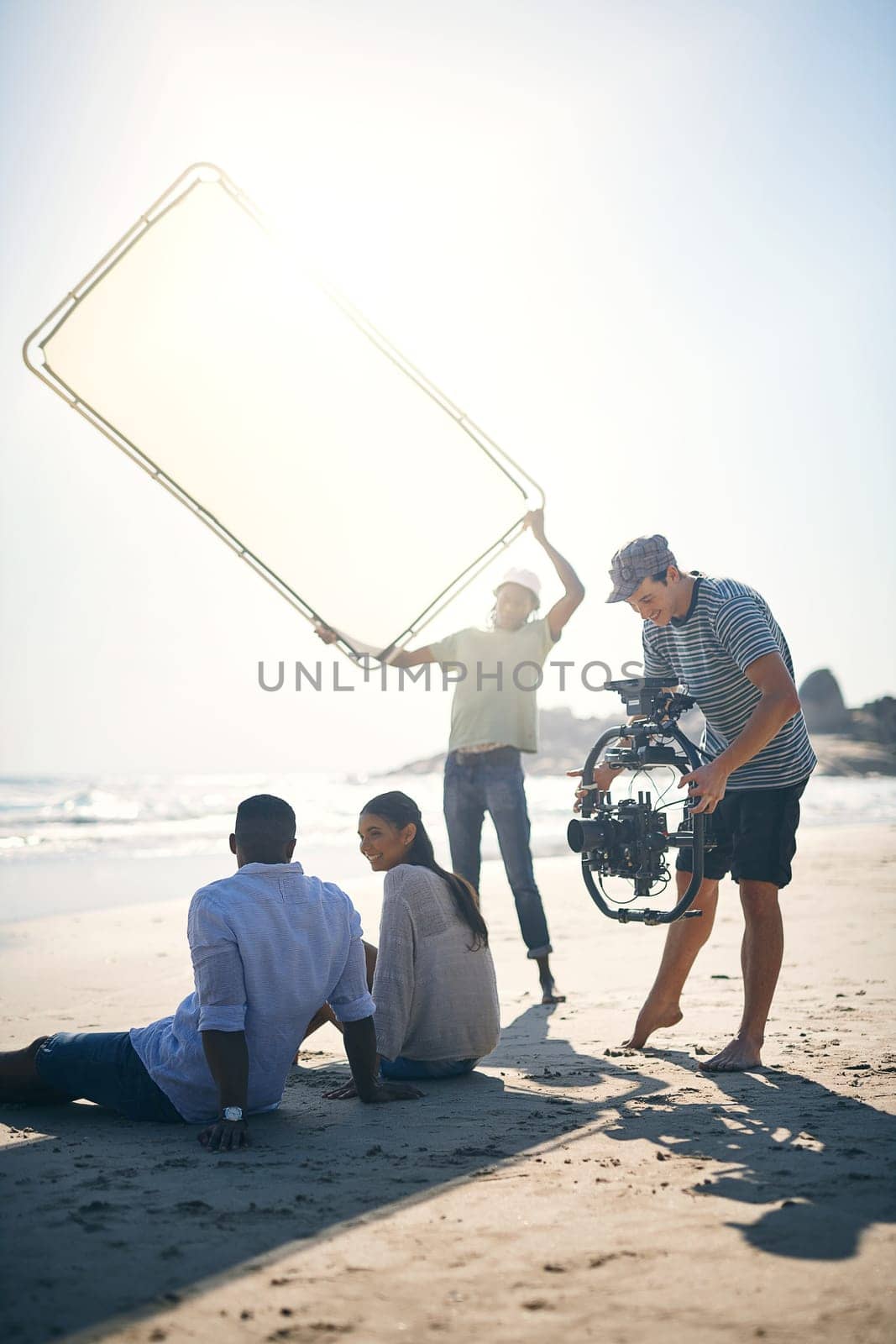 Bounce those sun rays. a man holding up a sun screen to dim the sun rays on two models that are being filmed by a videographer