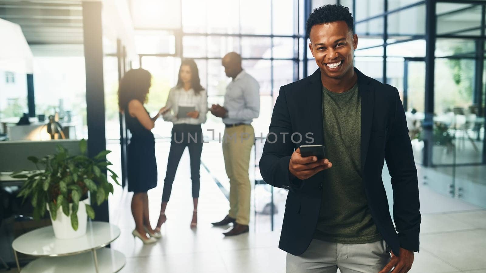 Technology helps me find all I need to operate business smoothly. Portrait of a young businessman using a cellphone in an office with his colleagues in the background. by YuriArcurs